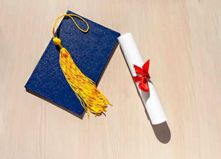 Yellow tassel from graduation cap, blue diploma and paper scroll tied with red ribbon with bow on beige wooden background, Flat lay, top view, mortarboardの写真素材