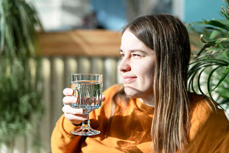 Profile of young pretty caucasian woman holding glass of drinking water in sunlight against backdrop of home plants, smiling, healthy lifestyleの写真素材
