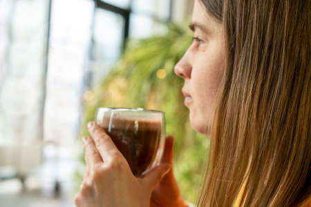 Young pretty Caucasian woman holding glass cup of coffee or cocoa and meditating looking out the window on background of green house plants, coffeetime, hot drinks, hot chocolateの写真素材