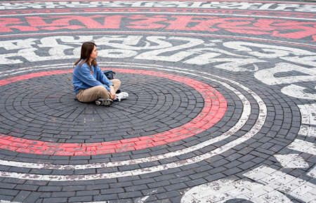 Young woman in blue sweatshirt alone sitting on street cobbled square in the form of circles and dreamily looking away, introvert, copy spaceの写真素材