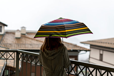 Young woman in khaki jacket from behind with umbrella in rainbow pattern in drops standing on the...