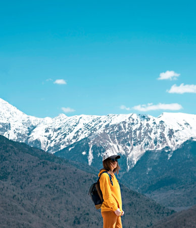 side view Young blond woman in yellow hoodie, cap, with backpack on background of snow capped...