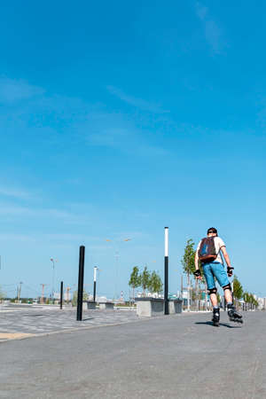Rear view of young man in blue shorts with backpack, in protective equipment riding on roller skates on the asphalt road in summer, roller blading, outdoor activity, healthy active lifestyleの写真素材