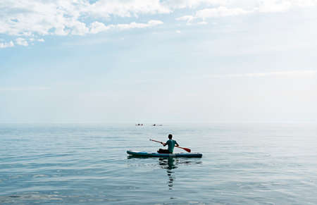 Young man from behind sitting on stand up paddle board on the sea on sunny summer day, active lifestyle, outdoor activityの写真素材