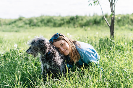Pretty young blond woman resting with fluffy gray dog on green grass among dandelions in summer, pet...