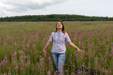 Happy Beautiful smiling blond young woman in a light purple shirt on a meadow of fireweed flowers and looking up at the sky at beginning of rain, copy spaceの写真素材