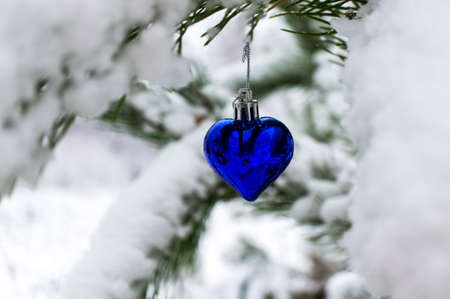 Christmas background with Christmas tree toy blue heart on snow covered pine branch in winter forest with copy space. New year holiday, Christmas celebration, greeting card. Merry Christmasの写真素材