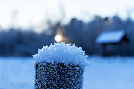 A snowy crystal cap on a wooden cut close-up and the setting sun at sunset against the background of a gazebo by the lake and forest. Abstract winter snowy background at sunsetの写真素材
