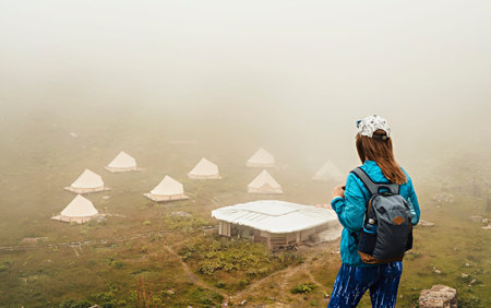 rear view of young woman in cap and blue sportswear with backpack hiker standing in green mountain...