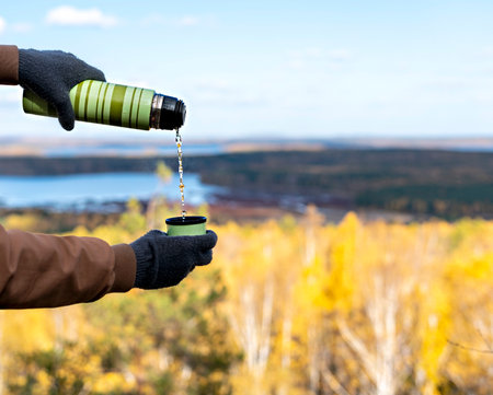 Male hands in warm gloves pour hot tea from flask on rock against the backdrop of beautiful autumn...