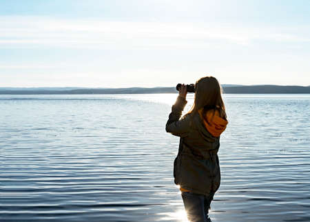 Young woman looking through binoculars at birds on the lake. Birdwatching, zoology, ecology. Research in nature, observation of animals Ornithology autumn bird migrationの写真素材