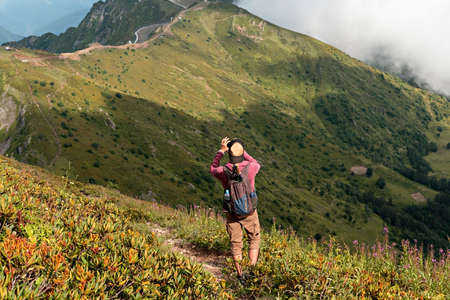 Young man with backpack hiking in Caucasus green mountains among the bushes of rhododendron and taking photo on smartphone, using modern technology active and healthy lifestyle, landscapeの写真素材