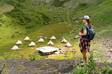 young woman in cap and plaid shirt with backpack hiker standing in green mountain valley against the backdrop of tent camping in fog, glamping, hiking selective focusの写真素材