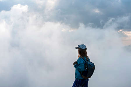 Young woman in cap and blue sporty clothes with backpack standing on the edge of mountain against background of white clouds, tourism hiking and active healthy lifestyle conceptの写真素材