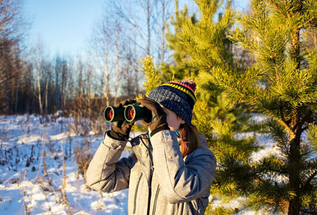 Young woman birdwatcher in winter clothes and knitted hat looking through binoculars in winter snowy...