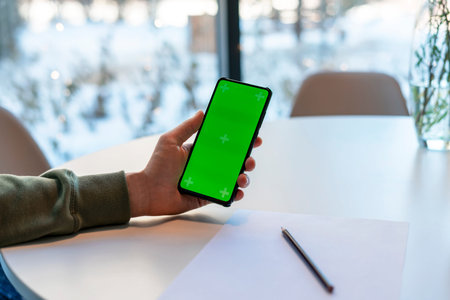 Man Holding Smartphone with Chroma Key Screen in cafe at white table, makes notes with pencil on...