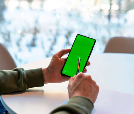 Man Holding Smartphone with Chroma Key Screen in cafe at white table, makes notes with pencil on paper , green screen man's hands business mock upの写真素材