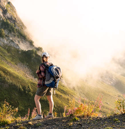 young woman in cap and plaid shirt with big backpack hiking in green mountains against clouds in summer healthy active lifestyle, outdoor activities, landscapeの写真素材
