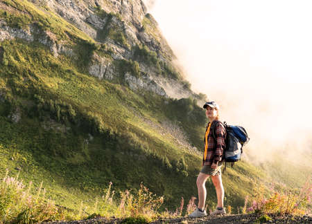 young woman in cap and plaid shirt with big backpack hiking in green mountains against clouds in summer healthy active lifestyle, outdoor activities, landscapeの写真素材