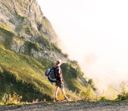 young woman in cap and plaid shirt with big backpack hiking in green mountains against clouds in...