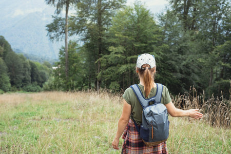 Rear view of a young woman in a cap with a backpack walking in the forest highlands in the summer...