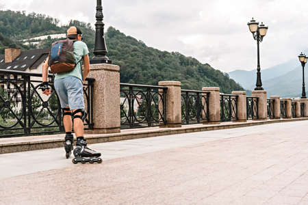 rear view of young man in blue clothes in protective equipment riding on roller skates along the embankment against the background of forest and mountains in summer, active lifestyle , selective focusの写真素材