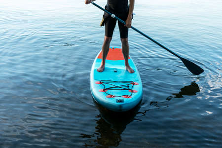 Man faceless in thermo suit paddling stand up paddleboard blue SUP on the water lake with paddle male legs active lifestyle summer activitiesの写真素材