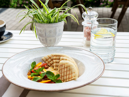 Hummus with tomatoes and herbs in a white plate with pita on the table and glass of water in cafe vegan foodの写真素材
