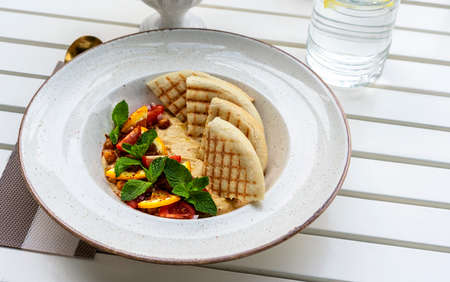 Hummus with tomatoes and herbs in a white plate with pita on the table and glass of water in cafe vegan foodの写真素材