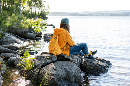 Young woman wearing cap, yellow hoodie with backpack sitting on rocks and looking at view of lake or...