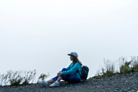 Young woman in cap and blue sporty clothes with backpack sitting on edge of mountain against white clouds and fog looking at view tourism hiking and active healthy lifestyle conceptの写真素材
