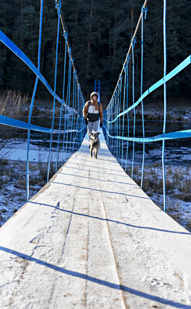 Young man with mixed breed fluffy dog walking on wooden bridge over river covered with first snow and ice in autumn or early winter Hiking and traveling with pets Active lifestyle dog adoptionの写真素材