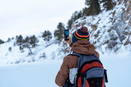 Rear view young man in brown warm clothes with backpack walking in snow among rocks and cliffs in winter looking at view taking photos with smartphone Active lifestyle hikingの写真素材