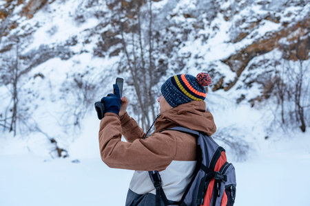 young man in brown warm clothes with backpack walking in snow among rocks and cliffs in winter...