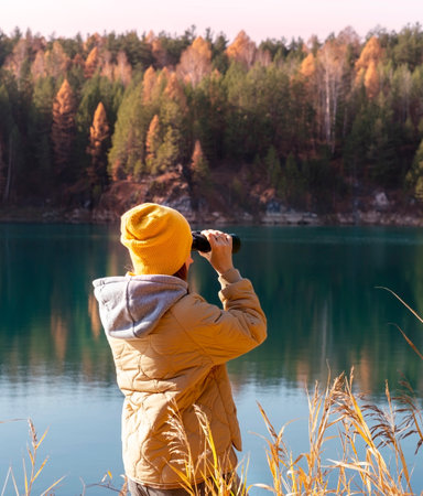 Young woman in yellow hat looking through binoculars at birds on lake against autumn forest Birdwatching, zoology, ecology. Research in nature, observation of animals Ornithologyの写真素材