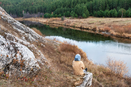 Young woman in beige jacket and hood sitting on rock and looking at the view of the river and forest in autumn seasonal neutral colors of nature tourism copy spaceの写真素材