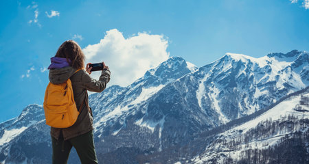 Young woman tourist with yellow backpack taking photo on smartphone of snow covered mountain range against blue sky modern technologyの写真素材