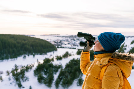 Young woman in yellow with binoculars looking at birds on mountain against winter snowy river and forest Birdwatching, zoology ecology Research in nature, observation of animals Ornithology copy spaceの写真素材