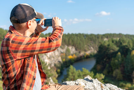 man in red plaid shirt sitting in rocks and taking photo on smartphone of mountains autumn forest and river landscape travel and tourism copy space using modern technology photographingの写真素材