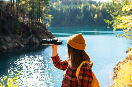 female traveler enjoys birdwatching by lake during autumn vacation, binoculars in hand, connecting with nature on an eco-friendly journeyの写真素材