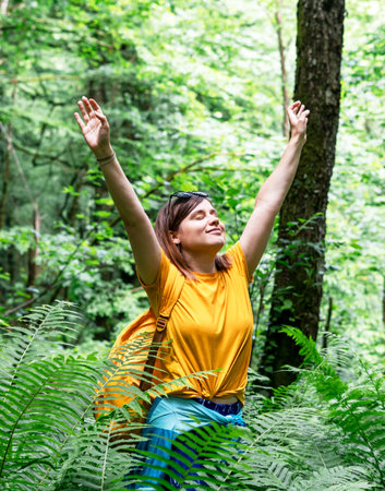 Happy young woman in yellow t-shirt with backpack raises her arms joyfully eyes closed in lush green forest, enjoys the beauty of nature, surrounded by ferns and tall trees , mental healthy lifestyleの写真素材