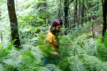 Young woman wearing a yellow shirt and backpack walking through a dense forest filled with green...