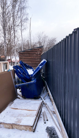 Blue trash bins stacked haphazardly in snow-covered alley. The bins are overflowing, indicating poor waste management. Snow dusts the ground and containers, accentuating the sceneの写真素材