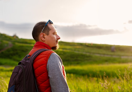 Man hiker wearing backpack and red vest stands outdoors, enjoying serene nature walk with the sun...