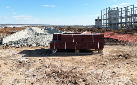 Construction site showcasing neatly stacked red tiles on pallets and a partially built structure in...