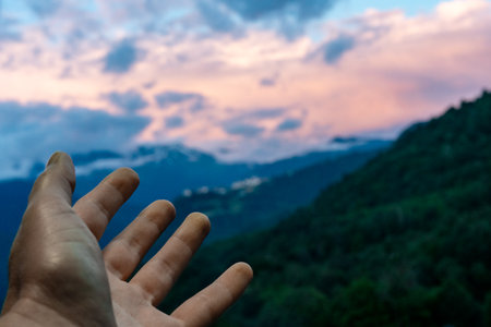 Outstretched hand reaching towards a distant landscape of mountains under cloudy sky at sunset, nature's beauty copy spaceの写真素材