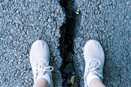 View of two feet in white sneakers standing at the edge of a deep crack in pavement. The scene highlights the condition of worn infrastructure and potential hazardsの写真素材