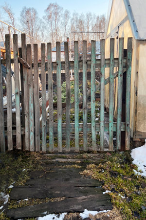 Rustic wooden garden gate leading to a backyard area during early spring. The scene captures remnants of snow melting on the ground, with bare trees in the backgroundの写真素材