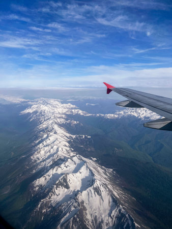 Snow-covered mountain range seen from airplane window with aircraft wing in view, blue sky , travel and transportationの写真素材