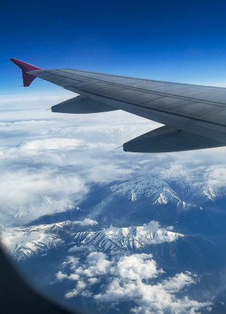View from airplane window showing wing , blue sky and snow-covered mountain range partially covered by clouds, transportation and travel conceptの写真素材